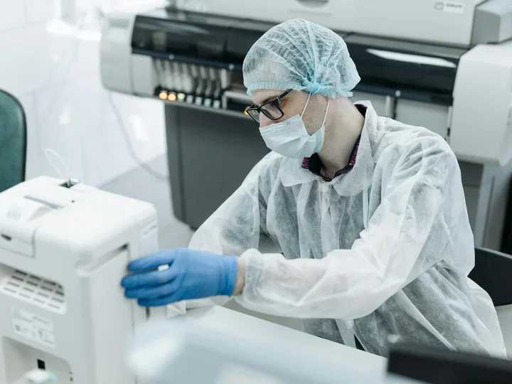 A person wearing protective clothing, a face mask, and gloves operates laboratory equipment in a clinical or research lab setting.